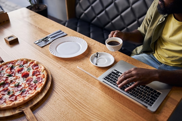 mężczyzna freelancer trzymający filiżankę kawy siedząc przy stole z laptopem i pizzą pepperoni