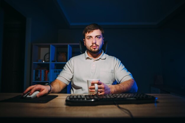 mężczyzna freelancer z brodą siedzi w nocy przy swoim biurku przy komputerze i pracuje z kubkiem napoju