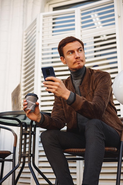 mężczyzna freelancer z filiżanką kawy przy użyciu smartfona i pracujący zdalnie w kawiarni na świeżym powietrzu