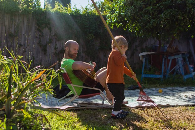 Zdjęcie mężczyzna gra na gitarze, patrząc na syna sprzątającego podwórko.
