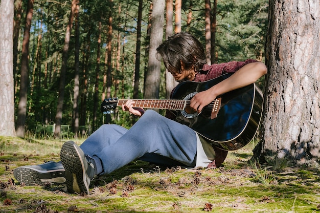 Mężczyzna gra na gitarze, siedząc w lesie w pobliżu drzewa. Na tle natury.