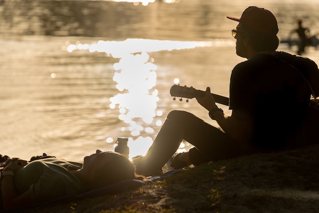 Mężczyzna grający na gitarze i kobieta leżąca na trawie nad brzegiem rzeki o zachodzie słońca w Noosa, Australia