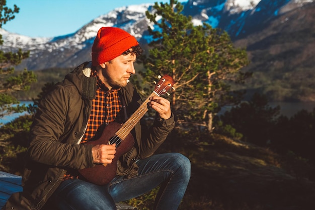 Mężczyzna grający na gitarze siedzący na drewnianym stole na tle gór, lasów i jezior