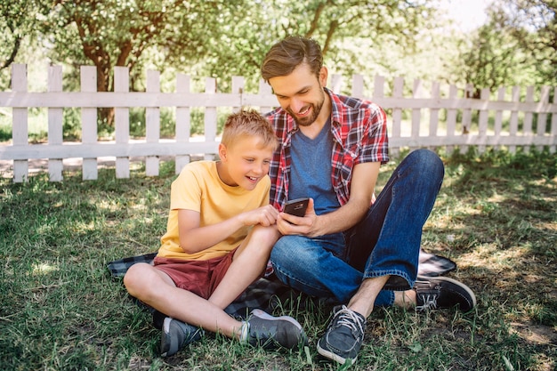 Mężczyzna i dziecko siedzą razem na trawie ze skrzyżowanymi nogami i patrząc na telefon