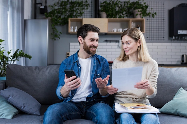 Mężczyzna i kobieta są szczęśliwi w domu dokonują płatności online za pomocą telefonu i aplikacji bankowej siedząc na kanapie i trzymając rachunki