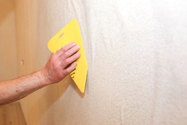 Mężczyzna instaluje tapetę za pomocą skrobaka. Poziomowanie ściany. Prace remontowo-remontowe remontowe w mieszkaniu. Renowacja w pomieszczeniu.