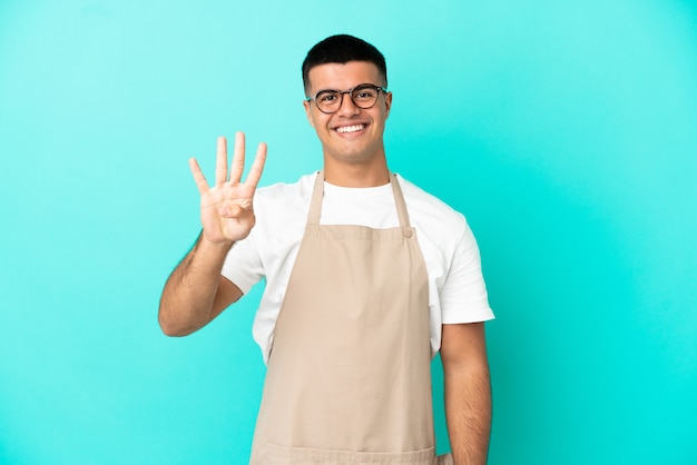 Mężczyzna kelner w restauracji na odosobnionym niebieskim tle szczęśliwy i liczący cztery palcami