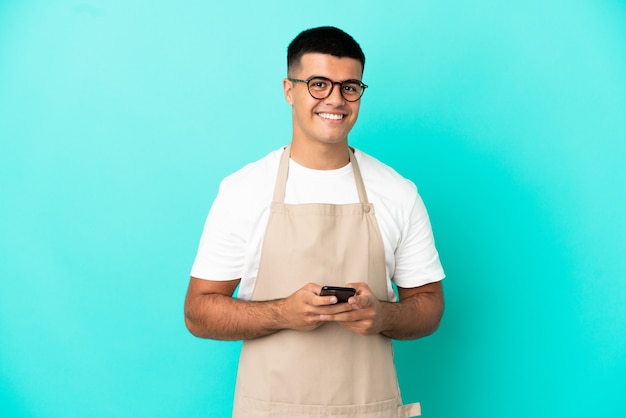 Mężczyzna kelner w restauracji na odosobnionym niebieskim tle wysyła wiadomość za pomocą telefonu komórkowego