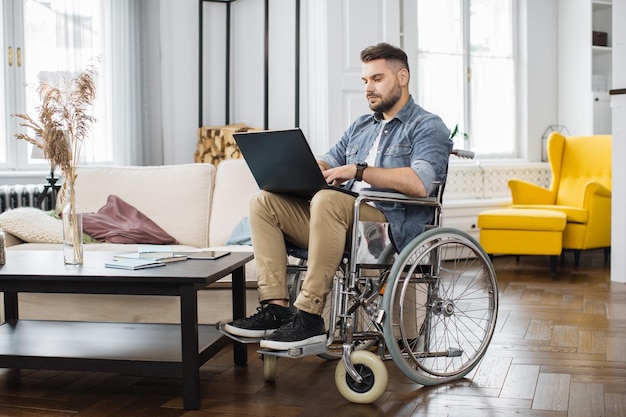 Mężczyzna korzystający z wózka inwalidzkiego, piszący na laptopie w salonie