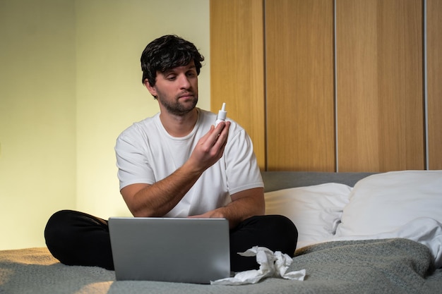 Mężczyzna, który czuje się chory, siedząc w domu na łóżku, kapie krople do nosa do zatkanego nosa Mężczyzna rozpyla lekarstwo na przeziębienie na katar i słucha lekarza za pomocą laptopa