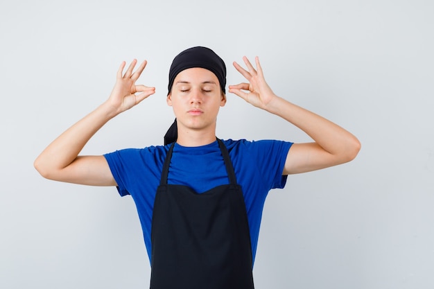 Zdjęcie mężczyzna kucharz nastolatek pokazując mudra znak w koszulce, fartuchu i patrząc spokojnie, widok z przodu.