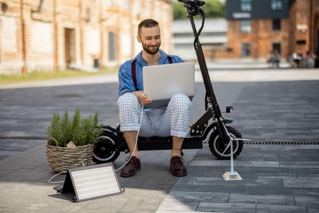 Mężczyzna ładuje laptopa panelami słonecznymi, siedząc na skuterze elektrycznym na zewnątrz