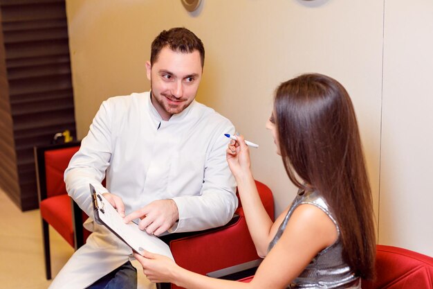 Mężczyzna lekarz i uśmiechnięta pacjentka, brunetka, siedzą w gabinecie lekarskim i wypełniają kwestionariusz