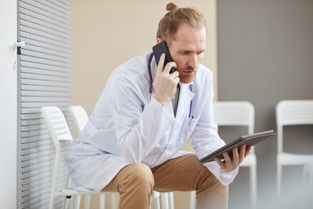 Mężczyzna lekarz rozmawia przez telefon komórkowy podczas korzystania z cyfrowego tabletu siedząc na krześle w szpitalu