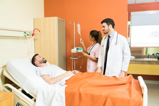 Mężczyzna lekarz rozmawia z pacjentem leżącym na łóżku szpitalnym z pracownicą opieki zdrowotnej dostosowującą natężenie przepływu kroplowego IV
