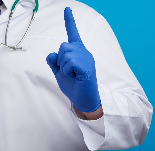 Mężczyzna lekarz w białym płaszczu, niebieskie rękawice medyczne pokazuje gest uwagi, palec wskazujący w górę