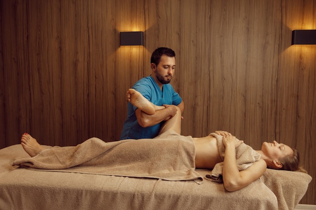 Mężczyzna masażysta ocierając nogę o szczupłą kobietę w ręczniku, profesjonalny masaż. Masaż i relaks, pielęgnacja ciała i skóry. Atrakcyjna pani w salonie spa