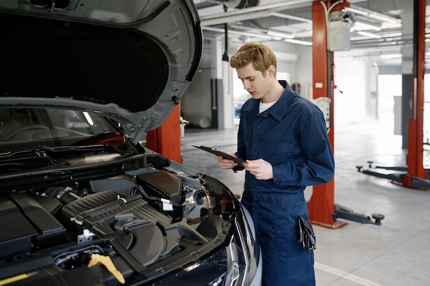 Mężczyzna mechanik stojący przed otwartym maską samochodu z kartką w ręku