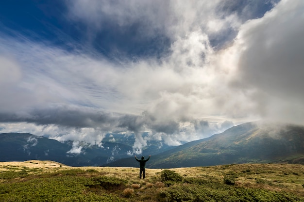 Mężczyzna na trawiastym wzgórzu z podniesionymi rękami na chmurnym niebieskim niebie i zielonych drzewiastych górach.