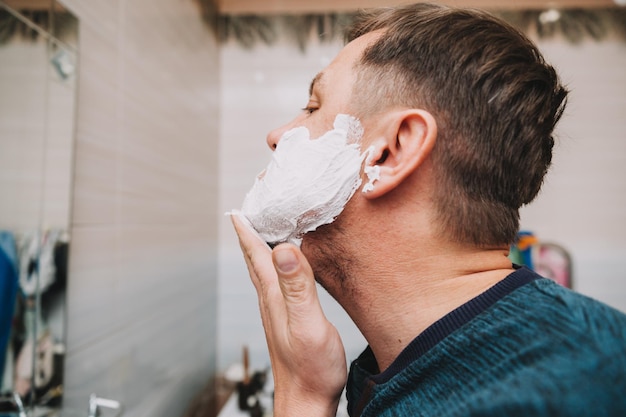 Mężczyzna nakłada piankę do golenia na twarz, aby golić się w łazience. higiena osobista.
