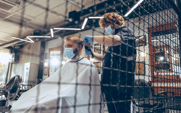 Mężczyzna obcina włosy u fryzjera w masce podczas pandemii koronawirusa. Profesjonalny fryzjer w rękawiczkach. Covid-19, koncepcja urody, samoopieki, stylu, opieki zdrowotnej i medycyny.