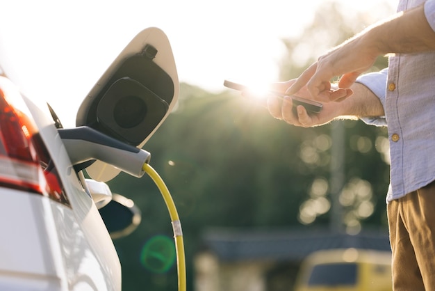 Mężczyzna odłączający przewód zasilający do samochodu elektrycznego za pomocą aplikacji na smartfonie biznesmen ładujący energię elektryczną