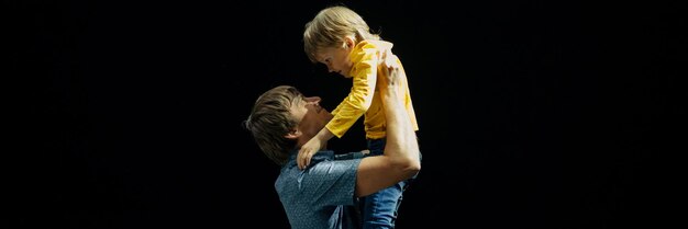Zdjęcie mężczyzna ojciec tata trzyma w ramionach małego chłopca syna w wieku sześciu lat i przytula szczęśliwe rodzinne rodzicielstwo ojcostwo i dzieciństwo w studio oświetlonym jasnym światłem i czarnym banerem tła
