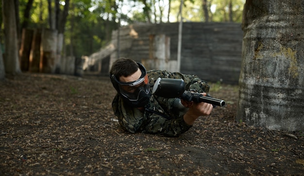 Mężczyzna paintballista z pistoletu strzela leżąc na ziemi, plac zabaw w lesie na tle. Sporty ekstremalne z bronią pneumatyczną i kulami lub markerami malarskimi, wojskowa gra zespołowa w plenerze