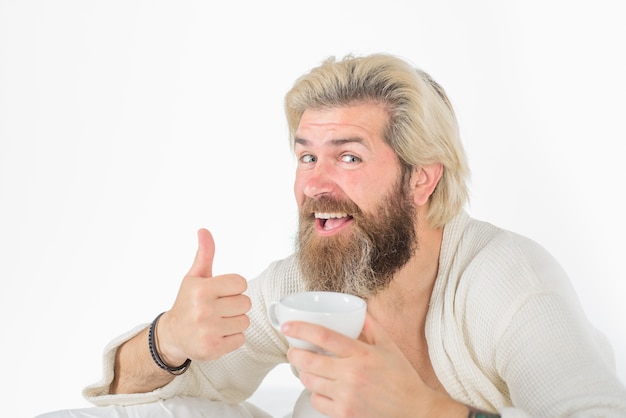 Mężczyzna pije kawę w łóżku śniadanie w łóżku brodaty mężczyzna w szlafroku pokazuje kciuk w porannej rutynie