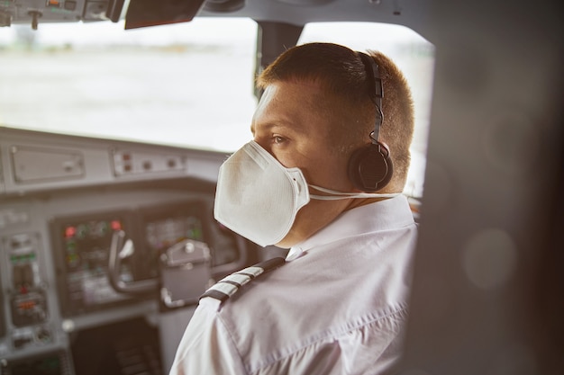 Mężczyzna Pilot W Kokpicie Samolotu Pasażerskiego. Widok Z Tyłu Europejskiego Człowieka Nosić Mundur, Maskę Medyczną I Słuchawki. Cywilne Lotnictwo Komercyjne. Koncepcja Podróży Lotniczej