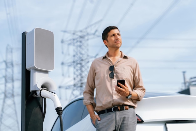 Mężczyzna płaci za energię elektryczną smartfonem podczas ładowania akumulatora samochodu elektrycznego na stacji ładowania podłączonej do wieży energetycznej jako przemysł elektryczny w celu przyjaznego dla środowiska wykorzystania samochodu