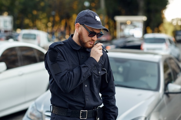 Mężczyzna policjant rozmawia przez radio na parkingu. Policjant w mundurze chroni prawo, rejestracja wykroczenia. Policjant pracuje nad ulicami miasta, kontrolą porządku i sprawiedliwości