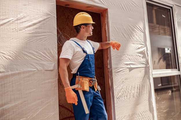 Mężczyzna pracownik budowlany noszący kask ochronny i kombinezon roboczy z paskiem narzędziowym, odwracając wzrok i uśmiechając się