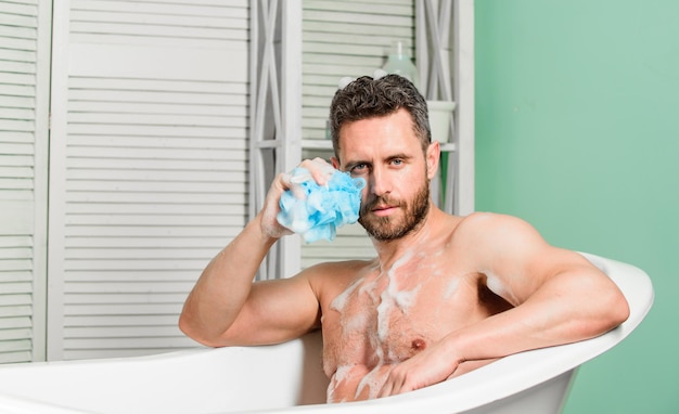 Mężczyzna przystojny muskularny facet relaksuje się w wannie Macho z gąbką kąpie się w domu Koncepcja metroseksualna Kąpiel z mydlinami Rozkoszowanie się gorącą kąpielą Rozpieszczanie i pielęgnacja