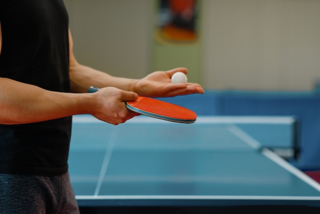 Mężczyzna ręce z rakietą do ping ponga i piłką, trening w pomieszczeniu. Mężczyzna w stroju sportowym stojący przy stole z siatką, trenujący w klubie tenisa stołowego