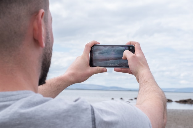 Mężczyzna Robi Zdjęcie Telefonem Komórkowym Na Plaży. Widok Na Góry W Oddali