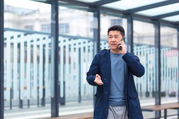 Mężczyzna rozmawiający przez telefon skupiony i poważny Azjata na przystanku transportu publicznego, który zatrzymuje biznesmena w pobliżu centrum biurowego