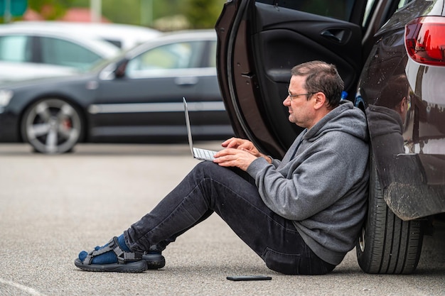 Mężczyzna siedzący na asfalcie samochodem i pracujący na laptopie, pracujący zdalnie, podróżujący