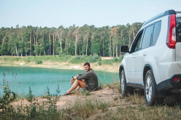 Mężczyzna siedzący w pobliżu białego samochodu typu suv na krawędzi, patrzący na jezioro z niebieską wodą