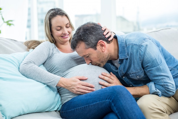 Mężczyzna siedzi na kanapie i całuje w ciąży brzuch kobiety