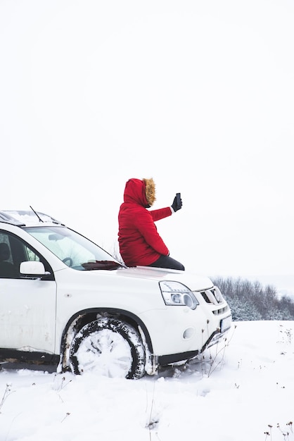 Mężczyzna siedzi na masce samochodu i robi zdjęcie pięknego zimowego widoku na swoim telefonie