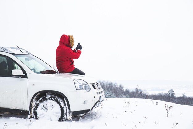 Mężczyzna siedzi na masce samochodu i robi zdjęcie pięknego zimowego widoku na swoim telefonie