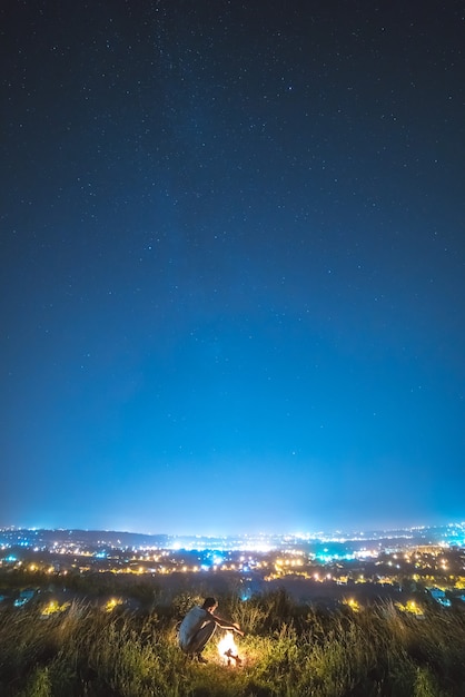 Mężczyzna siedzi przy ognisku na tle miejskich świateł. pora nocna