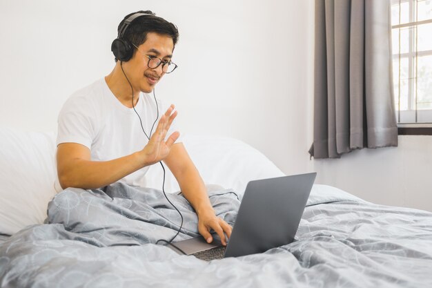 Mężczyzna siedzi w łóżku i macha rękę z hełmofonami ma wideo połączenie na laptopie