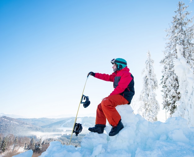 Mężczyzna snowboarder siedzi na szczycie śnieżnego wzgórza ze swoim snowboardem