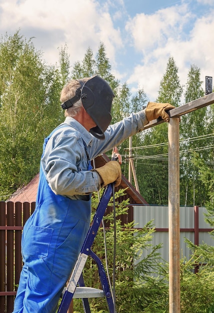 Mężczyzna Spawacz W Mundurze Budowlanym Maski Spawalniczej I Rękawicach Ochronnych Gotuje Metal Na Ulicznej Budowie Budowa Pergoli Pawilonu W Pobliżu Wiejskiego Domu W Letni Dzień