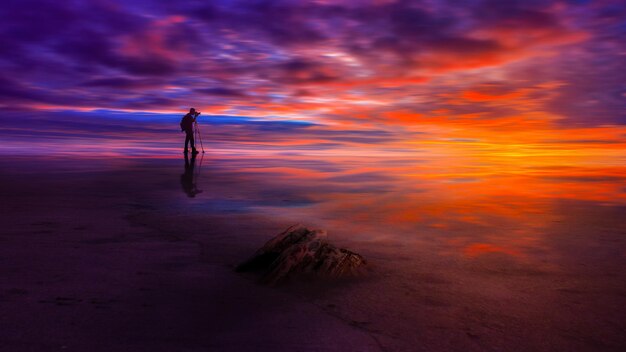 Mężczyzna stoi na plaży z dużą skałą na pierwszym planie, a niebo jest pomarańczowe i fioletowe.
