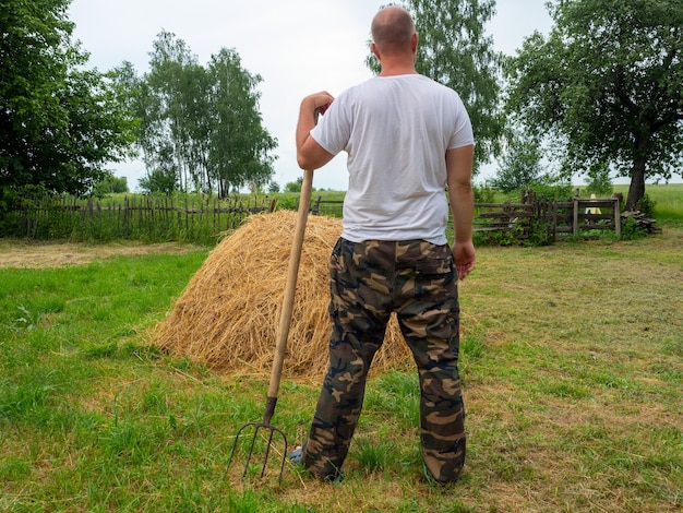 Zdjęcie mężczyzna stoi tyłem do kamery z widłami przed stogiem siana. wiejski krajobraz