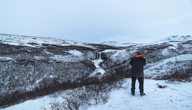 Mężczyzna stojący na całej długości, osłonięty od polarnego zimna, robiący zdjęcia wodospadu Svartifoss