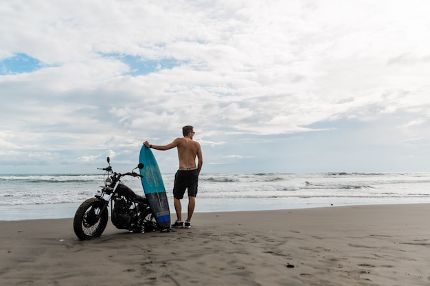 Mężczyzna szuka inspiracji, stojąc w pobliżu sportowego motocykla. trzymając w rękach deskę surfingową. Młody człowiek surfer korzystających z rekreacji w pobliżu oceanu.